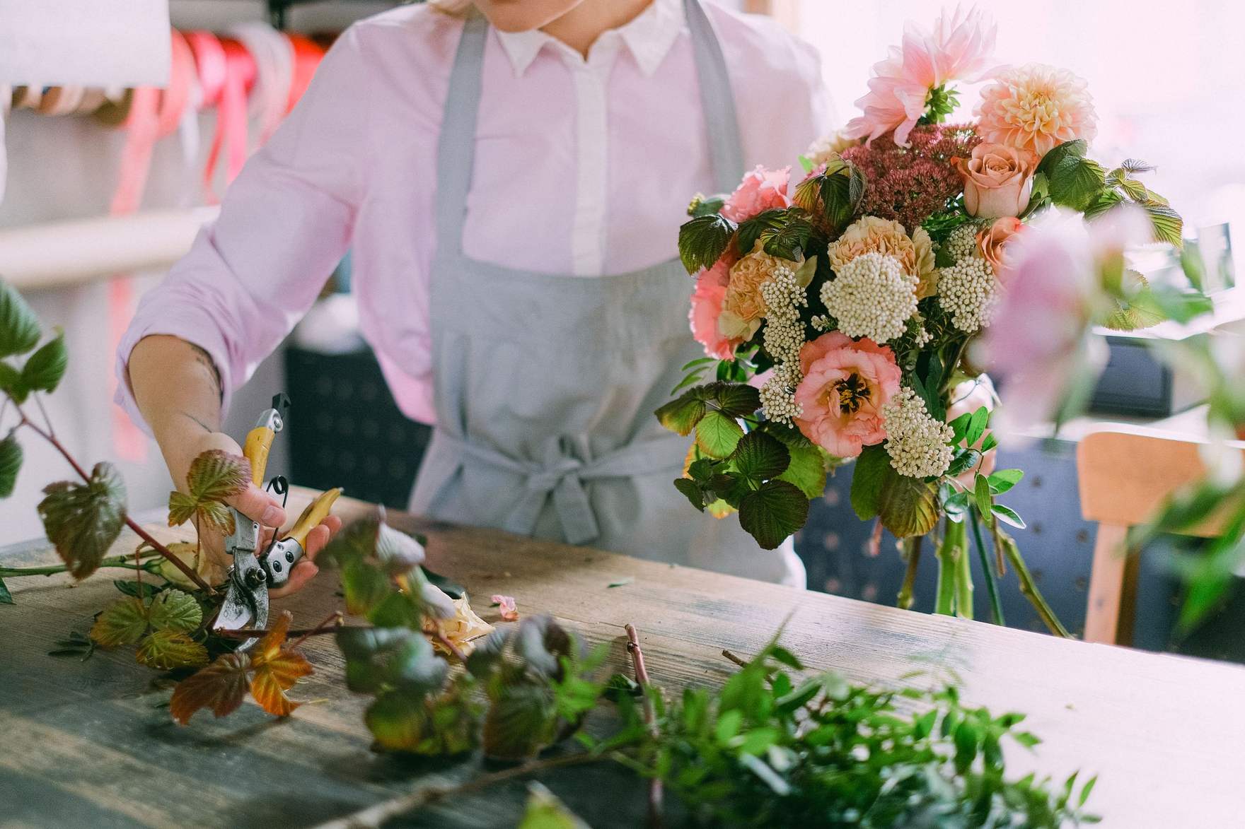 A florist in an apron trimming stems at a wooden bench beside a blush and green hand-tied bouquet.