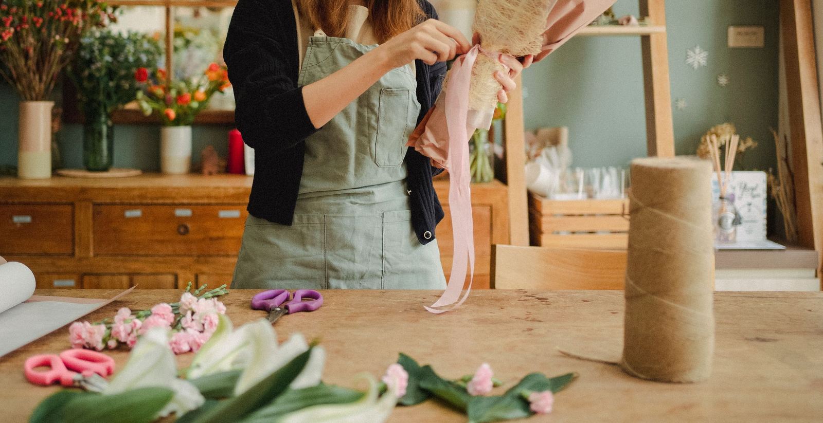A florist tying pink ribbon around a paper-wrapped bouquet on a workbench in a sage green studio.