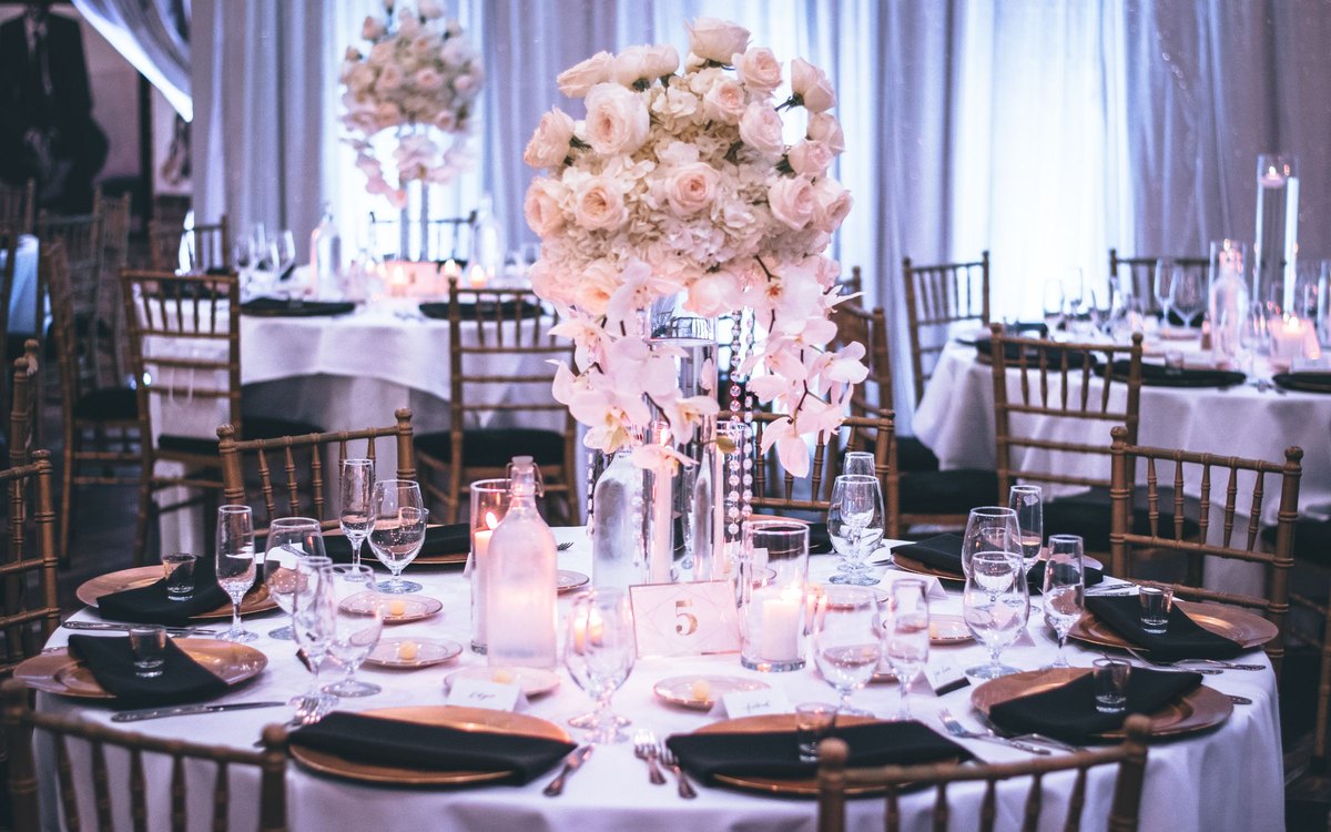 A wedding reception table set with a tall blush rose and orchid centerpiece, candles, and glassware.